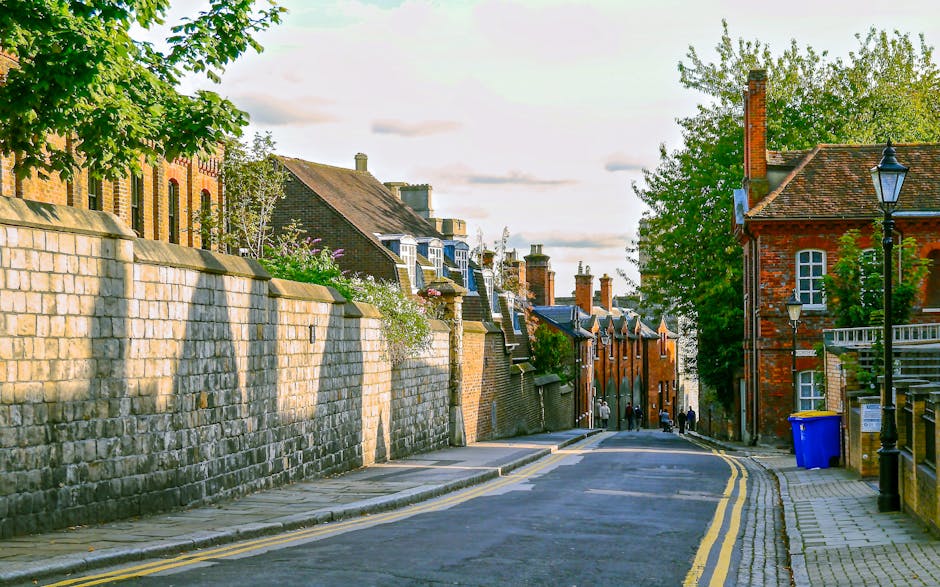 A narrow residential street with a paved road featuring double yellow lines on both sides, bounded by a high stone wall on the left and red brick buildings on the right. The stone wall has a slight slope and is topped with a row of greenery, while the brick buildings display traditional architecture with multiple chimney stacks and pitched roofs. Mature trees with dense foliage are visible along the street, casting shadows on the pavement. In the foreground, a blue recycling bin is positioned near a black lantern-style streetlight mounted on a pole beside the sidewalk. The scene is illuminated by soft, late afternoon sunlight, creating a warm atmosphere. This setting reflects a typical urban environment where house removals, furniture transport, or home relocation services by [COMPANY_NAME] could encounter access challenges due to the narrow road and surrounding architecture, emphasizing the importance of efficient moving logistics and careful handling of furniture and packing during the loading process for targeted removals in Kingston upon Thames.