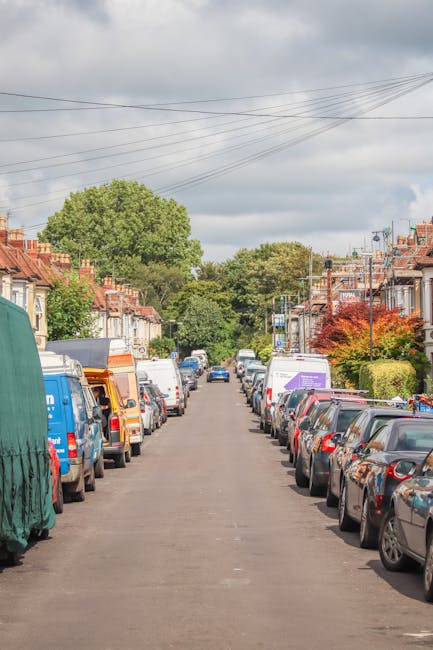 A narrow urban street in Kingston upon Thames with a slightly wet asphalt surface, flanked by multi-storey buildings on both sides. On the left, there are modern brick and glass commercial and residential buildings, while on the right, there are white and grey facades, possibly part of a mixed-use property. A white van is parked near the centre of the street, surrounded by a few small groups of pedestrians walking along the pavement. In the foreground, a moving trolley loaded with cardboard boxes, packing materials, and blankets for furniture transport is visible, positioned near the entrance of a property; the boxes are in various sizes and are wrapped with plastic or fabric for protection, indicating a home relocation or packing process managed by [COMPANY_NAME]. Behind the trolley, other packing materials and small pieces of furniture may be present, suggesting a careful loading process involved in the removals service. The scene is illuminated by natural daylight under a cloudy sky, capturing the typical environment for house removals and moving logistics in a busy city setting.