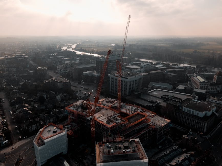 An aerial view of a busy urban construction site during daytime showing multiple orange tower cranes towering over a partially completed building with concrete floors and scaffolding. Surrounding the construction, there are various commercial and residential buildings, some with flat roofs, and a network of streets with parked cars. In the background, a river winds through the cityscape, with more buildings and green spaces visible in the distance under a slightly overcast sky. This scene captures the ongoing home relocation infrastructure development or commercial property expansion relevant to house removals and moving logistics, as seen in the context of professional removals services offered by manwithvankingstonuponthames.com.