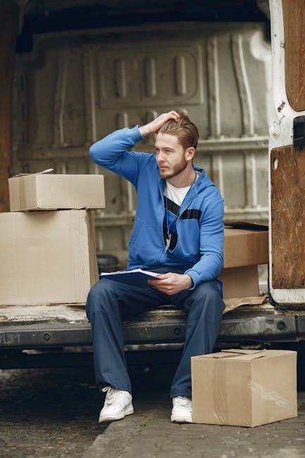 A young man with light brown hair, dressed in a blue jacket, dark trousers, and white sneakers, is seated on the edge of a van's loading area during a home relocation. He holds a clipboard in his lap and scratches his head, indicating a moment of pause or reflection. Surrounding him are several cardboard boxes of various sizes, some sealed with packing tape, placed inside the van and on the pavement. The interior of the van is visible behind him, showing a metallic, paneled space designed for moving furniture and household items. The scene captures the process of loading or unloading during a furniture transport operation, with the man likely coordinating or managing the packing and moving process, a service offered by companies like [COMPANY_NAME]. The lighting is natural, suggesting daytime, and the background includes part of the vehicle’s side and an outdoor setting, emphasizing a residential move within a suburban or urban environment.