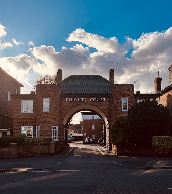 A brick residential building with an arched driveway entrance labeled 'KNIGHTS COURT', situated on a quiet street in daylight with a partly cloudy sky overhead. Visible on the property are several parked cars beneath the arch and around the driveway, with a man in casual clothing loading or unloading cardboard boxes wrapped in plastic and fabric moving blankets onto a trolley and into a van positioned outside the entrance. The scene suggests active furniture transport or home relocation involving multiple boxes and packing materials, aligned with local removals services by MAN WITH VAN KINGSTON UPON THAMES. The surrounding area features neatly maintained bushes and pathways, and the overall setting reflects a typical house removals process during a house move or commercial relocation with appropriate equipment, packaging, and vehicle access.