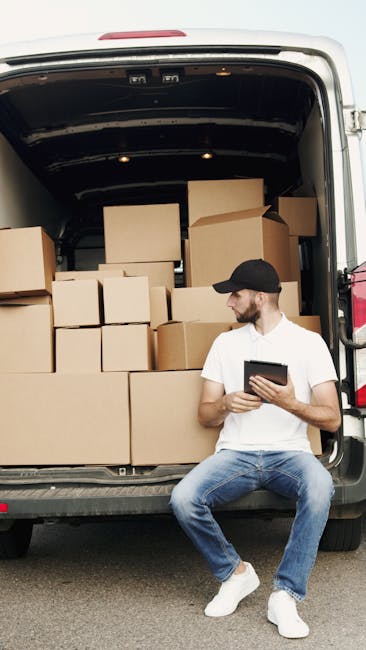 A man wearing a white t-shirt, light blue jeans, white sneakers, and a black cap is sitting on the edge of an open van with its rear doors fully open. Inside the van, there are multiple cardboard boxes of various sizes, some stacked neatly and others arranged to allow efficient packing for a house relocation. The boxes are sealed with packing tape, and some are wrapped in plastic for protection. The man is holding a tablet device, likely coordinating or checking a packing list, while resting during the loading process. The van is parked on a paved surface, and the lighting appears natural, indicating daytime. This scene depicts a professional furniture transport and packing stage related to house removal services, supported by [COMPANY_NAME], in preparation for a house move or furniture transport arrangement in Kingston as part of a local moving operation associated with the Bentall Centre shop relocation for Kingston businesses.
