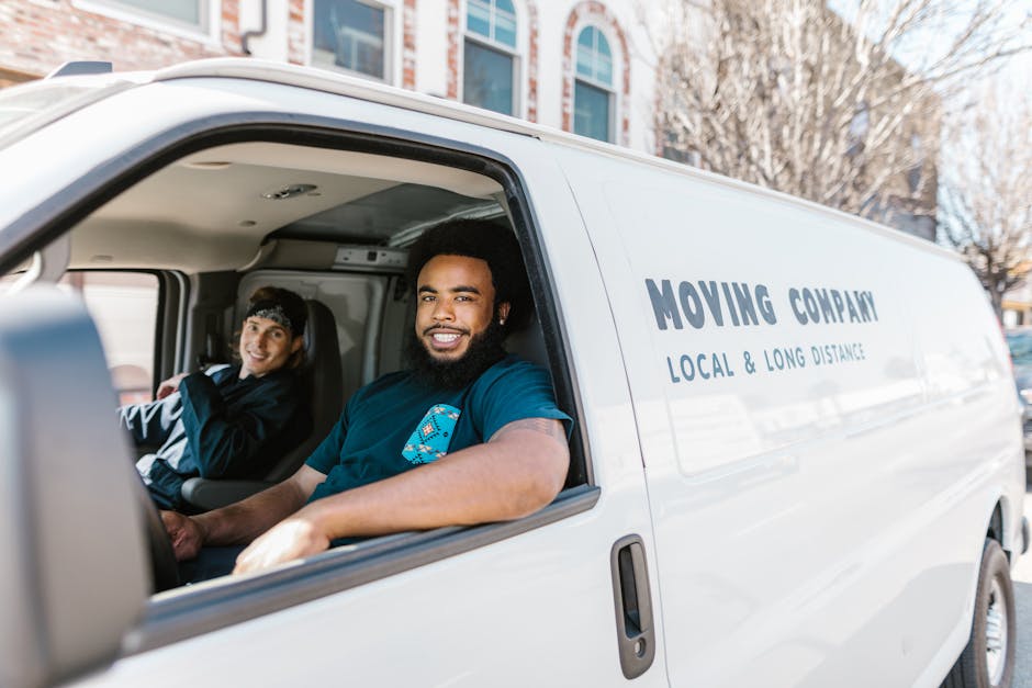 Inside a white moving van parked outdoors, two male movers are visible through the open driver's side window. The man in the passenger seat, a Black male with curly hair and a full beard, is smiling, wearing a blue t-shirt, and appears to be ready for home relocation or furniture transport tasks. The driver, a male with light skin and dark hair, is seated behind the steering wheel and also smiling, dressed in a dark jacket. The van has professional branding with the words 'MOVING COMPANY' and 'LOCAL & LONG DISTANCE' printed on the side panel. The background shows a residential area with a multi-story building featuring large windows and trees with bare branches, suggesting a cool or transitional season. The scene captures a moment during a packing and moving process, emphasizing the loading or transportation phase associated with house removals, handled by [COMPANY_NAME], which specializes in removals services.
