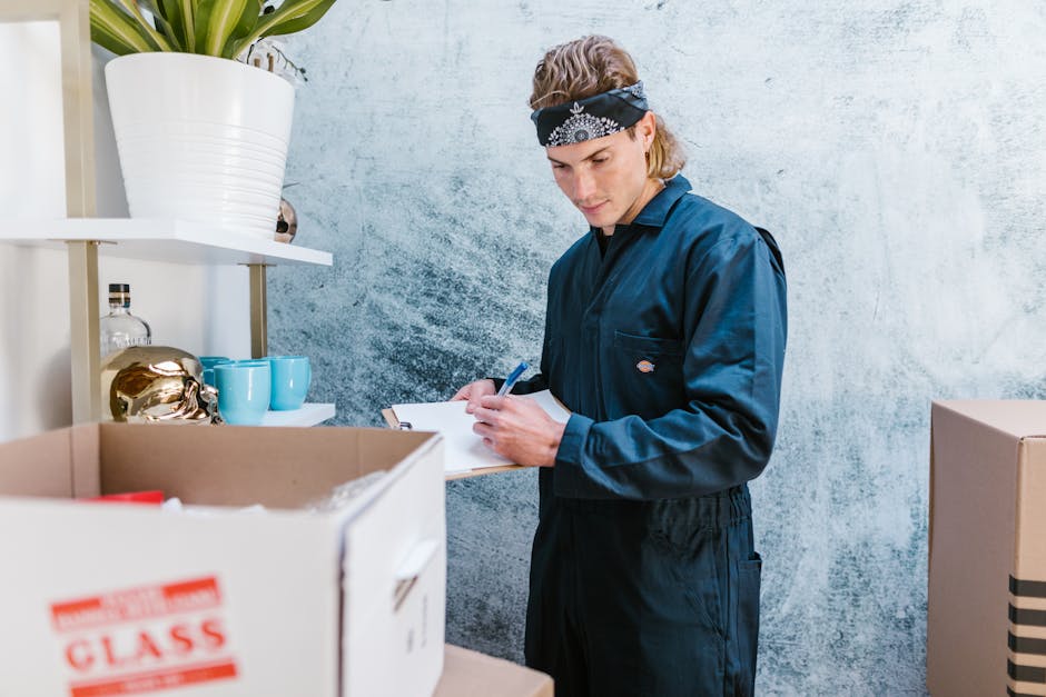 A young man with light brown hair, wearing a blue work uniform and a black bandana with white patterns, is inside a room, standing next to a cardboard box filled with various packing materials. He is holding a clipboard and a pen, appearing to write or check inventory related to house removals. On a nearby white shelf, there are decorative items, including a large white ceramic pot with a green potted plant and some glass bottles, as well as light blue cups. The room has a textured, light-colored wall, and the scene captures a moment of packing or organizing items in preparation for a house relocation, reflecting the processes involved in furniture transport and packing for a move, as detailed by the removals services of manwithvankingstonuponthames.com.
