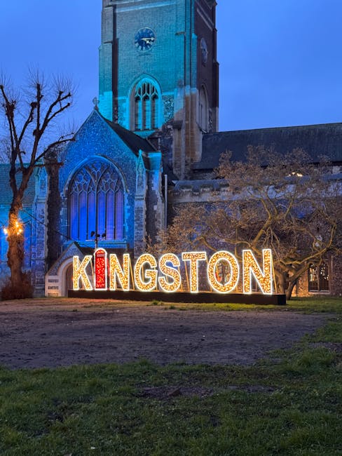 A nighttime view of Kingston's historic church with a tall clock tower illuminated in blue light, showing large stained glass windows on its facade. In front of the church, there is a brightly lit sign spelling 'KINGSTON' with red and white bulbs, placed on the ground amidst leafless trees. The surrounding area includes a paved pathway and patches of grass, suggesting an outdoor public space often associated with home relocation and moving services like those provided by [COMPANY_NAME], helping clients with furniture transport and packing during house removals. The scene emphasizes the importance of planning loading and unloading areas when organizing home moves, with clear visibility of the church's architecture and the decorative signage conveying a welcoming community atmosphere.