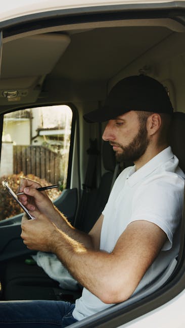 A man with a beard, wearing a black cap and a white T-shirt, is seated inside a moving van’s cab, looking at his smartphone. The vehicle’s interior is visible, including the ceiling and side window, through which a residential garden with a wooden fence and some plants can be seen. The man appears to be preparing for a home relocation, possibly coordinating logistics or confirming arrangements during a furniture transport process. The scene reflects the careful planning involved in packing and moving, with the man potentially overseeing the loading process or communicating with the removals team, which would be typical for a professional house removal service like those offered by manwithvankingstonuponthames.com.