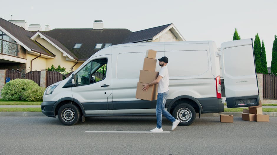 A man wearing casual clothing, including a white t-shirt, jeans, and a black cap, is carrying three cardboard boxes stacked together towards a white commercial van parked on a residential street in Kingston upon Thames. The van is positioned parallel to the curb with its side door open, indicating active loading or unloading during a home relocation or furniture transport process. Several additional cardboard boxes are placed on the pavement near the rear of the van, and the man appears to be carefully handling the boxes as part of a packing and moving operation. In the background, there are modern houses with pitched roofs, balcony features, and well-maintained gardens, along with tall green trees, under a clear sky. This scene illustrates a typical loading process during house removals, capturing the logistics involved in home relocation services provided by companies like manwithvankingstonuponthames.com as referenced in the Kingston Station removals guide for KT1 flats.