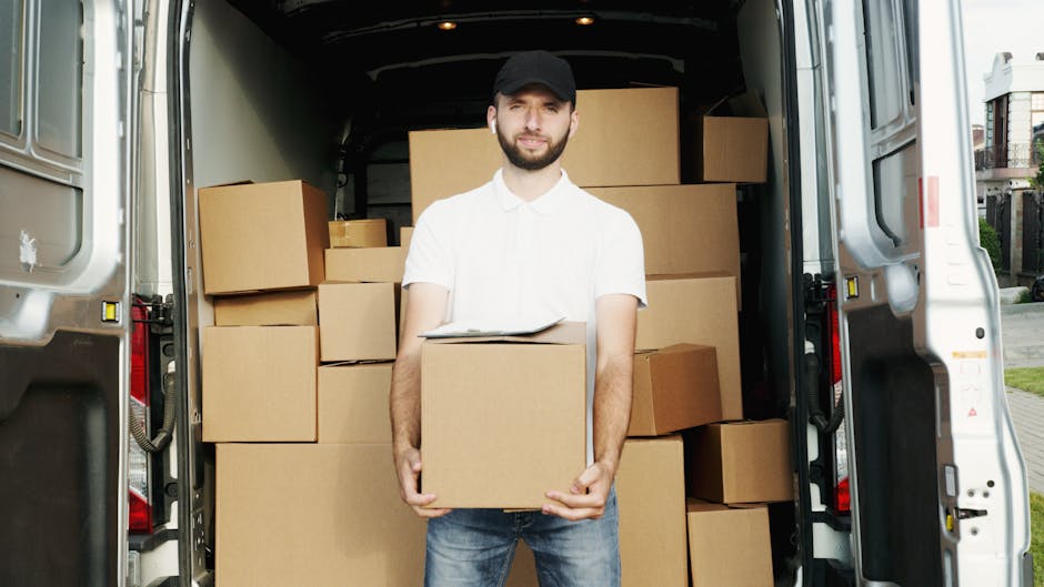 A man wearing a white polo shirt, jeans, and a black cap stands inside the open rear of a moving van, holding a cardboard box. The van's interior is filled with several other cardboard boxes, some stacked and others placed on the floor, ready for home relocation. The boxes are made of brown corrugated cardboard, some wrapped in clear plastic or packing tape. The van is parked outside a residential area on a paved street, with a row of houses visible in the background. The lighting indicates it is daytime, and the scene depicts the packing and loading process involved in furniture transport and house removals, with [COMPANY_NAME]'s services supporting efficient packing and moving logistics for customers in Kingston and the surrounding areas.