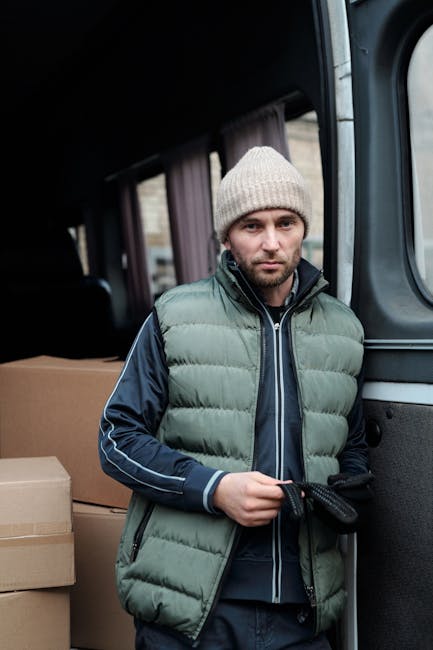A young man with a serious expression stands next to a black moving van, holding a black strap used for securing items during a home relocation. He is dressed in a grey knitted beanie, a green padded vest over a dark blue jacket with white stripes on the sleeves, and dark gloves. Inside the van, visible behind him, are several cardboard boxes placed on the floor, ready for loading or unloading. The background shows a window and part of the van’s interior, indicating an indoor or driveway setting during a furniture transport or packing and moving process, typical of professional removals services offered by companies like manwithvankingstonuponthames.com.