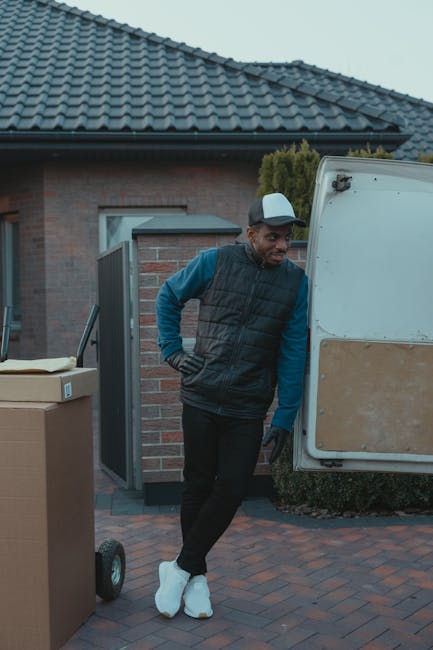 A man wearing a white polo shirt, jeans, and a black cap stands inside the open rear of a moving van, holding a cardboard box. The van's interior is filled with several other cardboard boxes, some stacked and others placed on the floor, ready for home relocation. The boxes are made of brown corrugated cardboard, some wrapped in clear plastic or packing tape. The van is parked outside a residential area on a paved street, with a row of houses visible in the background. The lighting indicates it is daytime, and the scene depicts the packing and loading process involved in furniture transport and house removals, with [COMPANY_NAME]'s services supporting efficient packing and moving logistics for customers in Kingston and the surrounding areas.