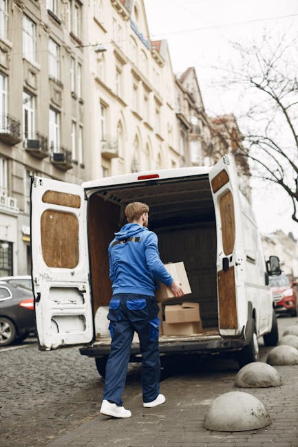 A man wearing a blue jacket and trousers is loading cardboard boxes into the open rear doors of a white commercial van parked on a cobblestone street in Kingston upon Thames. The van's interior is visible, showing wooden panels and packaging materials, indicating preparation for a home relocation or furniture transport. The street is lined with multi-storey residential buildings featuring classic architectural details, including balconies and large windows. Several cars are parked along the street, and leafless trees are visible in the background, suggesting a colder season. The trolleys and straps associated with moving and packing are not present in this specific image, but the scene consistently reflects a professional packing and loading process, supporting the services of [COMPANY_NAME] for last-minute Kingston same-day removals and emergency moves.