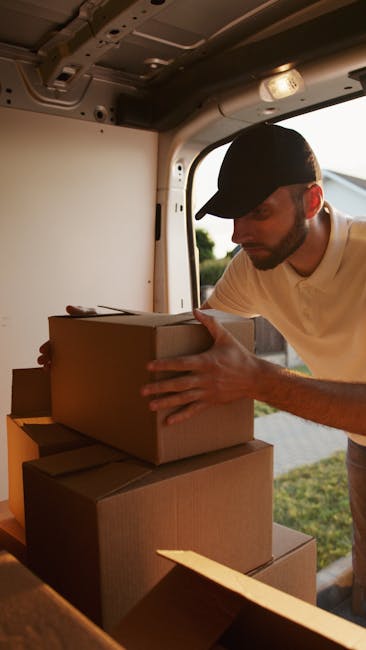 A man wearing a blue jacket and trousers is loading cardboard boxes into the open rear doors of a white commercial van parked on a cobblestone street in Kingston upon Thames. The van's interior is visible, showing wooden panels and packaging materials, indicating preparation for a home relocation or furniture transport. The street is lined with multi-storey residential buildings featuring classic architectural details, including balconies and large windows. Several cars are parked along the street, and leafless trees are visible in the background, suggesting a colder season. The trolleys and straps associated with moving and packing are not present in this specific image, but the scene consistently reflects a professional packing and loading process, supporting the services of [COMPANY_NAME] for last-minute Kingston same-day removals and emergency moves.