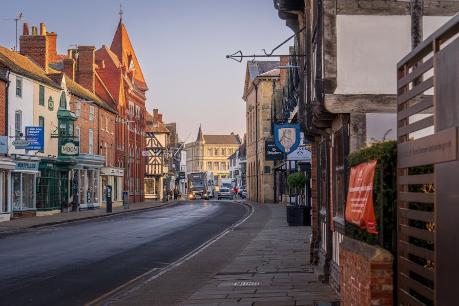 A quiet street scene in Kingston upon Thames showing a row of mixed architectural style buildings, including brick and timber-framed facades, some with signs advertising local businesses. The paved si