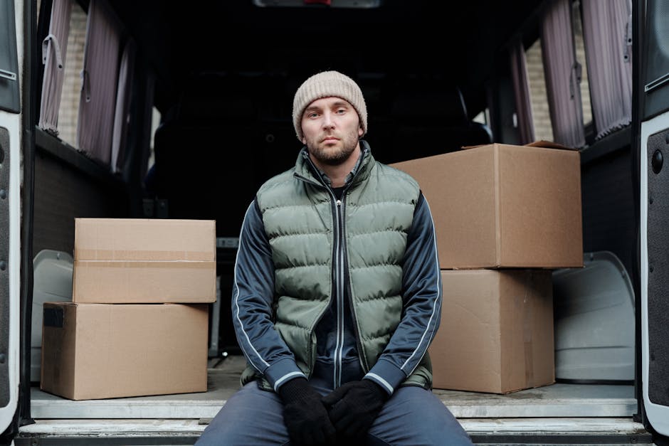 A man with a serious expression is seated on the edge of an open van with sliding doors, surrounded by cardboard boxes of various sizes that are stacked and arranged for a home relocation. The man is wearing a grey beanie, a green padded vest over a navy blue jacket with white stripes on the sleeves, and black gloves. The interior of the van shows a loading area with protective blankets and straps used for securing furniture during transportation. The background indicates an outdoor setting, likely near a residential property. This scene illustrates the logistics involved in packing and loading items for furniture transport and house removals, typical of professional moving services such as those offered by [COMPANY_NAME].