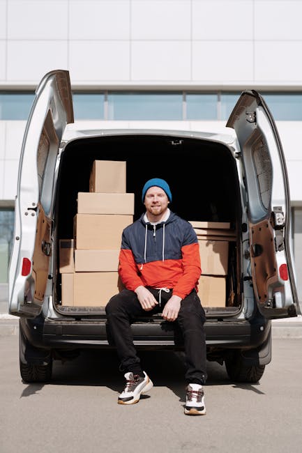 A man with a serious expression is seated on the edge of an open van with sliding doors, surrounded by cardboard boxes of various sizes that are stacked and arranged for a home relocation. The man is wearing a grey beanie, a green padded vest over a navy blue jacket with white stripes on the sleeves, and black gloves. The interior of the van shows a loading area with protective blankets and straps used for securing furniture during transportation. The background indicates an outdoor setting, likely near a residential property. This scene illustrates the logistics involved in packing and loading items for furniture transport and house removals, typical of professional moving services such as those offered by [COMPANY_NAME].